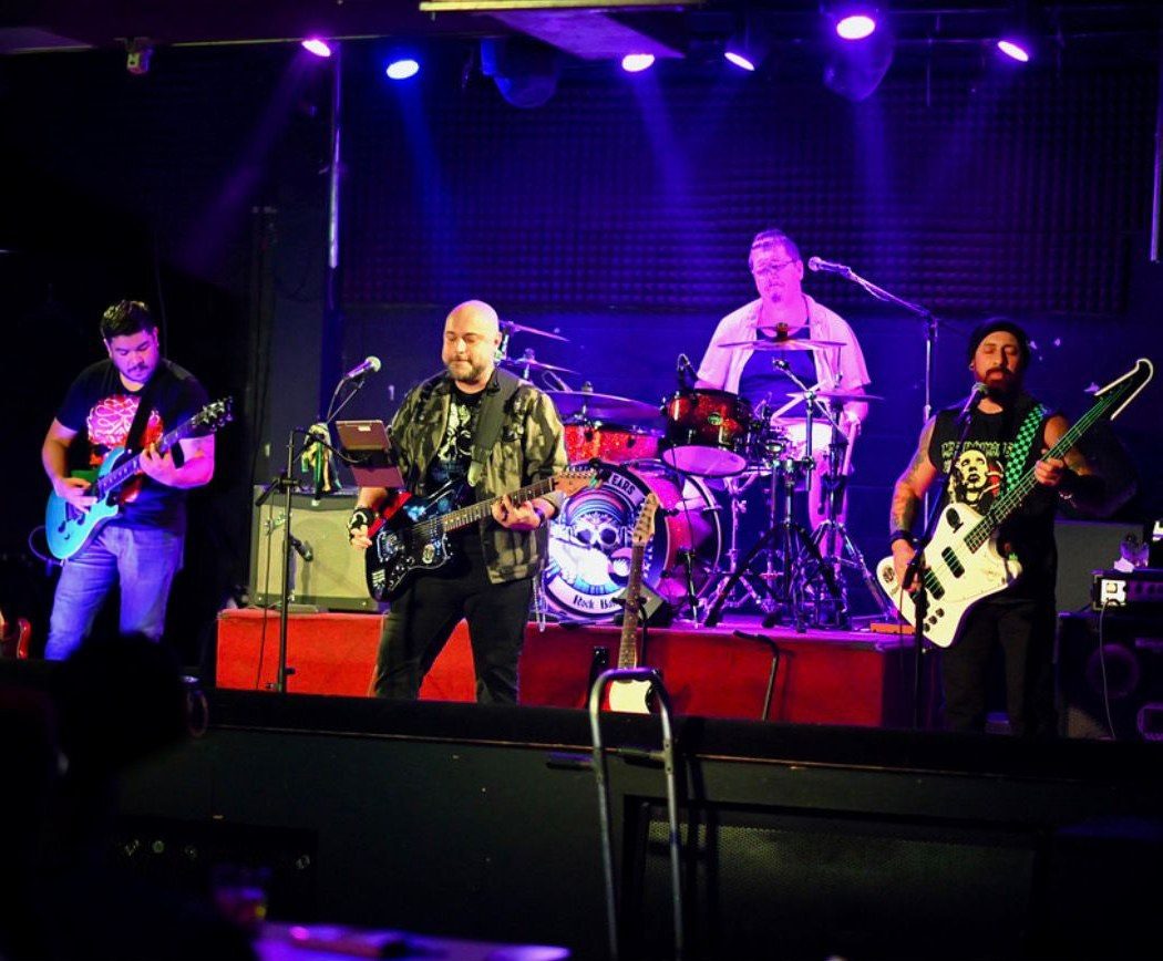 The full band on stage under purple lights: guitarist, Carlos, drummer and bassist