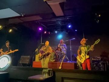 The band on a dark club stage lit by purple and blue lights